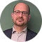 Professional headshot of a man with glasses and a beard, wearing a suit and standing against a green background.