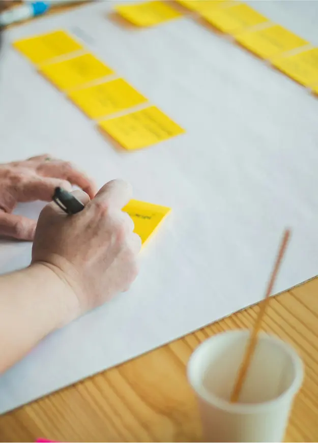 A hand writing on a yellow sticky note, surrounded by more sticky notes and a cup with a paintbrush.