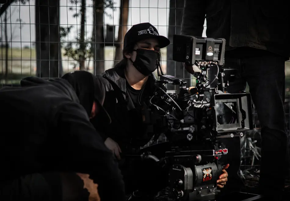 Filmmaker wearing a mask operates a camera on a film set, with another crew member nearby.