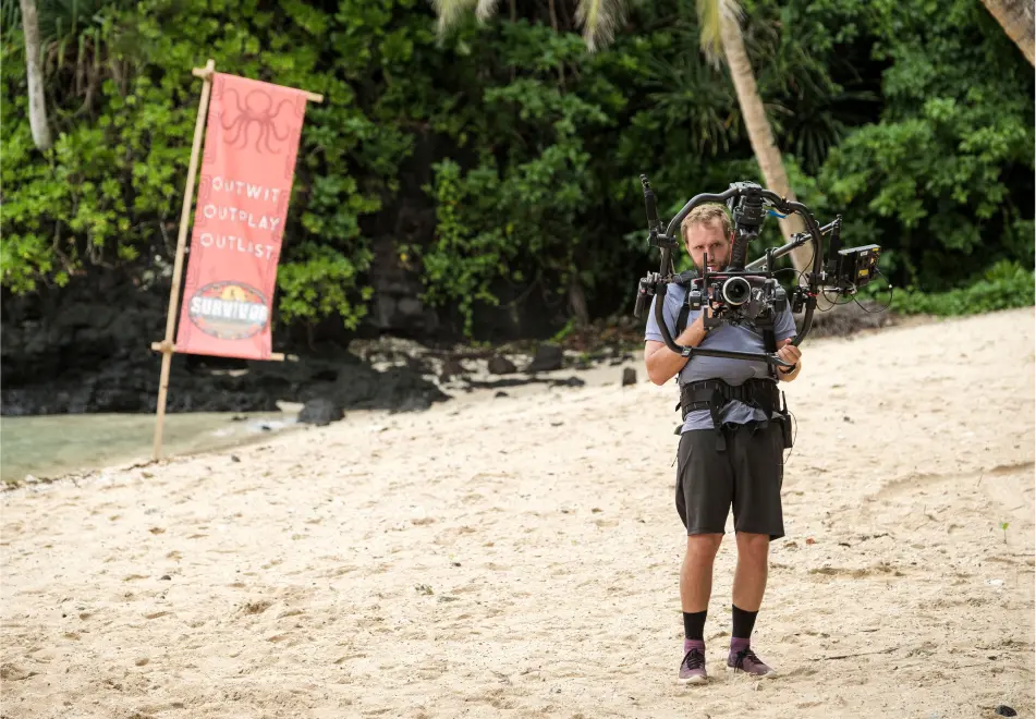 A cameraman stands on a sandy beach while holding a camera rig, with greenery and a banner in the background.